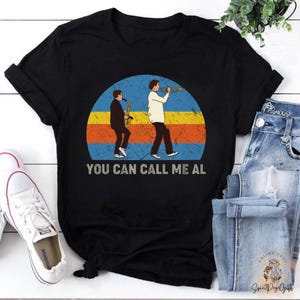 May include: Black t-shirt with a retro design featuring two musicians, one with a saxophone and the other with a trumpet, set against a blue, orange, and yellow background. The text "YOU CAN CALL ME AL" is printed below the graphic.