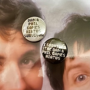 May include: Two round buttons with black text on a white background. The button on the left says "Dan & Phil Games Hiatus Survivor". The button on the right says "I Survived the Dan & Phil Games Hiatus".