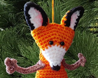 May include: A crocheted orange fox ornament with black and white details. The fox has a white belly and a black nose. The ornament is hanging from a green pine tree.