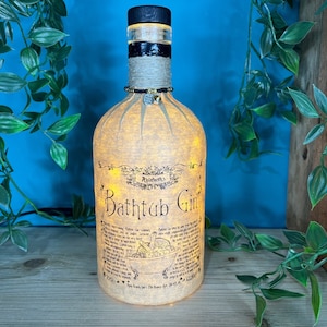 May include: A decorative bottle lamp made from a repurposed gin bottle. The bottle features a black wooden stopper, a black and white band, and a label that reads "Bathtub Gin." The bottle is illuminated with warm white lights.