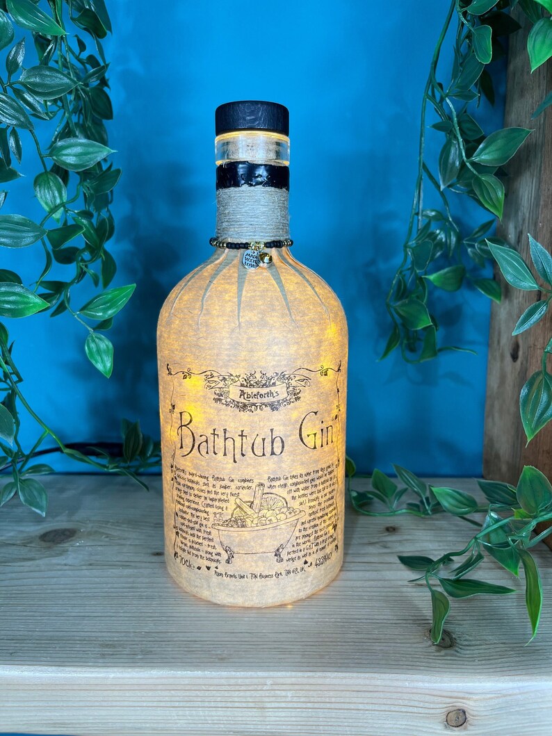 May include: A decorative bottle lamp made from a repurposed gin bottle. The bottle features a black wooden stopper, a black and white band, and a label that reads "Bathtub Gin." The bottle is illuminated with warm white lights.