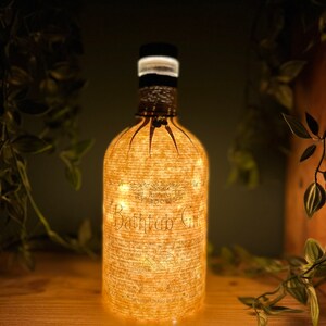 May include: A decorative bottle with the words "Bathtub Gin" illuminated by small lights. The bottle has a black cap and is wrapped in a textured, light-colored material. The bottle is sitting on a wooden surface.