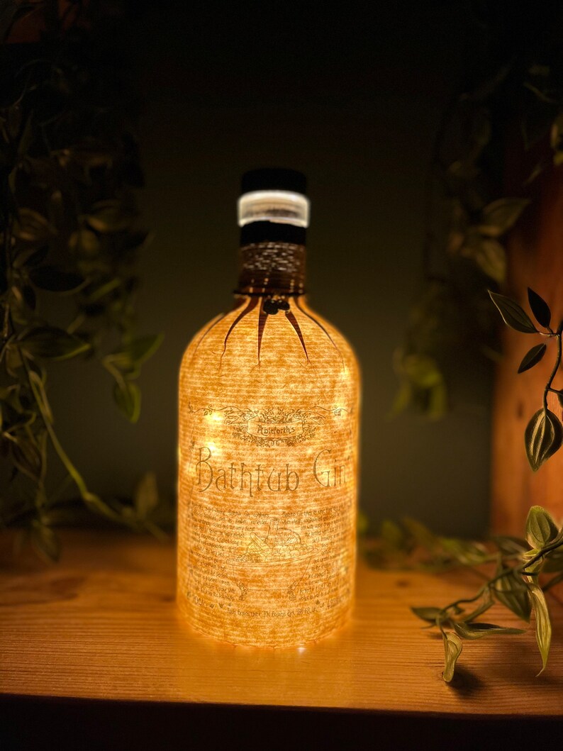 May include: A decorative bottle with the words "Bathtub Gin" illuminated by small lights. The bottle has a black cap and is wrapped in a textured, light-colored material. The bottle is sitting on a wooden surface.