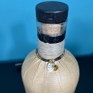 May include: A decorative bottle with a black stopper, wrapped in brown paper and twine. A beaded charm with a silver tag that says "Made with Love" and a yellow gem hangs around the neck of the bottle. The bottle is against a blue background.