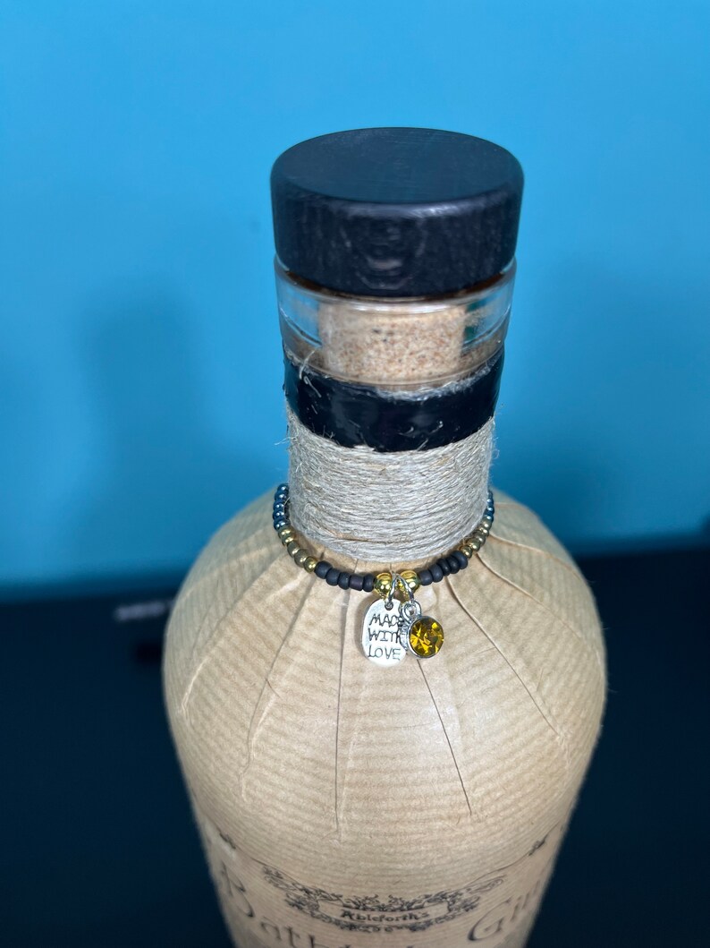 May include: A decorative bottle with a black stopper, wrapped in brown paper and twine. A beaded charm with a silver tag that says "Made with Love" and a yellow gem hangs around the neck of the bottle. The bottle is against a blue background.