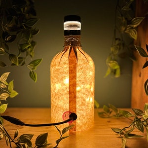 May include: A decorative bottle-shaped lamp with a warm, inviting glow. The lamp is wrapped in a textured, light-colored material, with a string of lights inside. The top is black with a white ring. A black cord extends from the base.