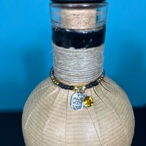 May include: A decorative bottle with a black stopper and a cork. The bottle is wrapped in twine and has a beaded charm with a silver tag that says "MADE WITH LOVE" and a yellow gem.
