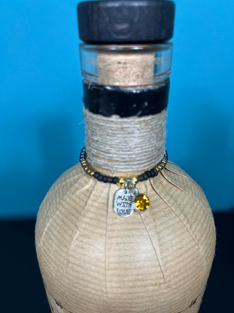 May include: A decorative bottle with a black stopper and a cork. The bottle is wrapped in twine and has a beaded charm with a silver tag that says "MADE WITH LOVE" and a yellow gem.