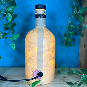 May include: A repurposed bottle lamp with a black cap and a beige paper shade. The bottle is lit from within, creating a warm glow. The lamp has a black power cord and a label that reads "ATOM SUPPLIES LTD".