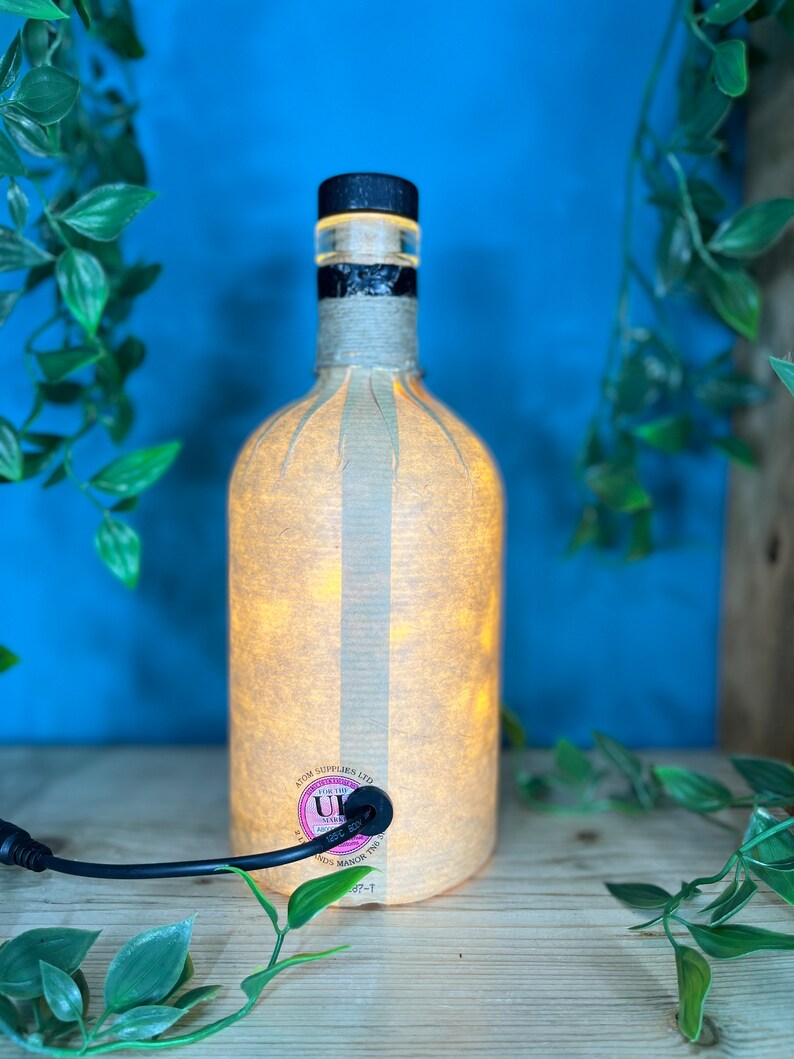 May include: A repurposed bottle lamp with a black cap and a beige paper shade. The bottle is lit from within, creating a warm glow. The lamp has a black power cord and a label that reads "ATOM SUPPLIES LTD".