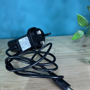 May include: A black AC adapter with a three-prong plug and a coiled black cable. The adapter has a small white label with text. The cable ends in a DC connector. The background is a light wood surface with a blue wall.