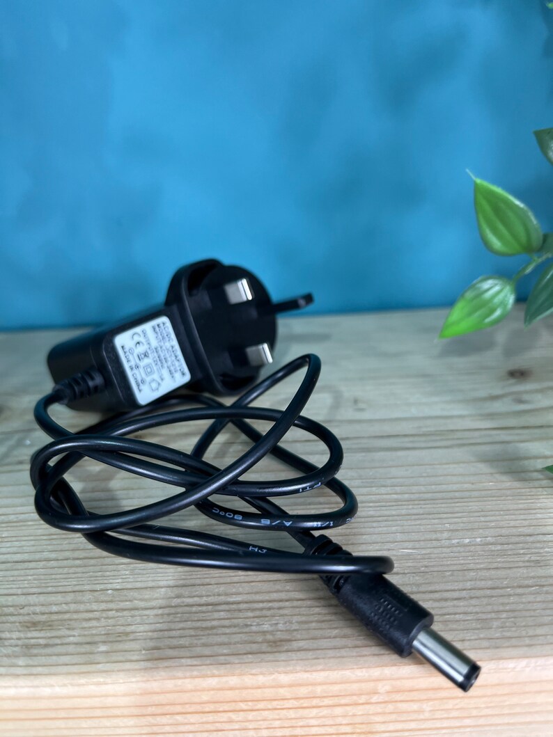 May include: A black AC adapter with a three-prong plug and a coiled black cable. The adapter has a small white label with text. The cable ends in a DC connector. The background is a light wood surface with a blue wall.