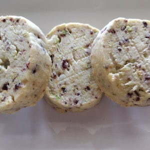 May include: Three round cookies with a light brown color and flecks of green and dark brown throughout. The cookies are arranged in a row on a white surface.