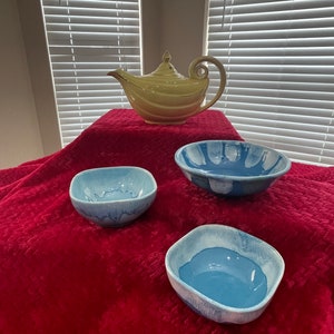 May include: A yellow teapot with a decorative design sits on a red tablecloth with three blue and white ceramic bowls.