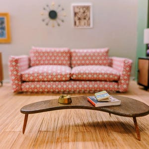 May include: A miniature living room scene featuring a pink and white patterned sofa, a dark brown kidney-shaped wooden coffee table, a small gold-toned bowl, and two tiny books. The room includes a wall clock, framed art, and side tables.