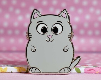 May include: A gray enamel pin featuring a cartoon cat with large eyes and a pink nose. The cat is sitting on its hind legs with its paws in front of it. The pin is a cute and whimsical design.
