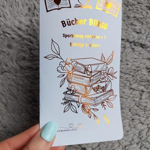 Może przedstawiać: Biała karta z literami i ilustracjami w kolorze złotym. Na karcie widnieje napis "Bücher Bingo" oraz "Sparbetrag 400 euro +? Beträge 1-6 Euro". Karta zawiera również złotą ilustrację stosu książek z kwiatami i motylami.