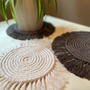 May include: Three macrame coasters in shades of white and brown are displayed on a white surface. The coasters have a circular design with a fringed edge. A potted plant sits on one of the brown coasters.
