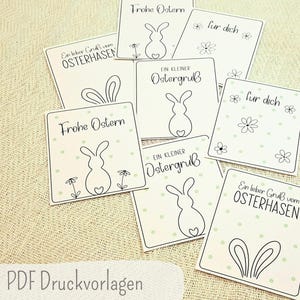 Könnte beinhalten: Eine Ansammlung weißer Karten mit Oster-Illustrationen und Text. Jede Karte zeigt eine schwarze Umrisszeichnung eines Hasen, Blumen und Text auf Deutsch. Die Karten liegen auf einer strukturierten, hellen Oberfläche.