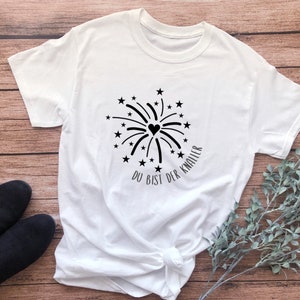 May include: White t-shirt with a black firework design and the text "Du bist der Knaller" in German.