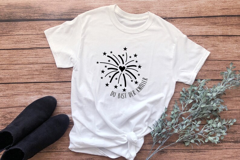 May include: White t-shirt with a black firework design and the text "Du bist der Knaller" in German.