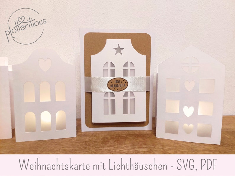 Op de afbeelding: Een witte kaart met een uitgesneden ontwerp van een huis met ramen en een ster bovenop. De kaart wordt van achteren verlicht, waardoor een gloei-effect ontstaat. De tekst "Frohe Weihnachten" is zichtbaar op de kaart. De kaart maakt deel uit van een set van drie kaarten met verschillende huisontwerpen.