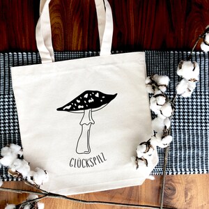 May include: A white canvas tote bag with black and white illustration of a mushroom and the text "Gl&uuml;ckspilz". The bag is lying on a wooden surface with a black and white checkered fabric and a cotton branch.