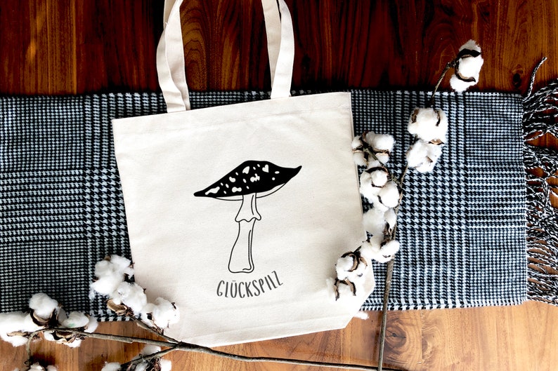 May include: A white canvas tote bag with black and white illustration of a mushroom and the text "Gl&uuml;ckspilz". The bag is lying on a wooden surface with a black and white checkered fabric and a cotton branch.