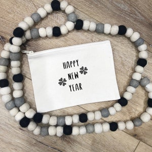 May include: A white zippered pouch with the text "Happy New Year" and two four-leaf clover symbols printed in black. The pouch is surrounded by a garland of white, gray, and black felt balls.