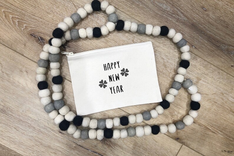 May include: A white zippered pouch with the text "Happy New Year" and two four-leaf clover symbols printed in black. The pouch is surrounded by a garland of white, gray, and black felt balls.