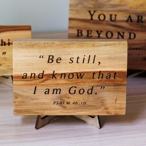 May include: Three rectangular wooden signs with natural grain patterns, each resting on a small wooden stand. The signs feature black engraved text: "Do everything in Love," "Be still, and know that I am God," and "YOU ARE LOVED BEYOND MEASURE."
