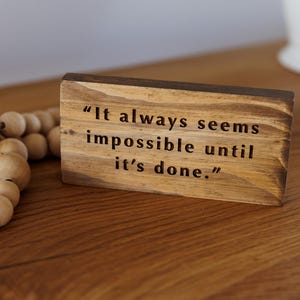 May include: A rectangular wooden block with a natural brown finish featuring the black engraved text "It always seems impossible until it's done." A strand of light-colored wooden beads is positioned to the left of the wooden sign.