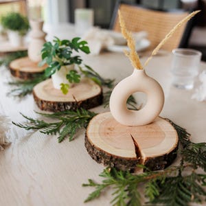May include: A table setting with three wooden slices, each with a small vase. The vases are white and have different shapes. The table is decorated with greenery.