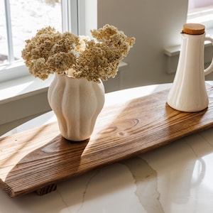 May include: A rectangular wooden riser with a dark brown grain finish and four small feet. On top sits a white ceramic vase with a scalloped texture filled with dried cream-colored flowers and a white ceramic pitcher with a wooden lid.