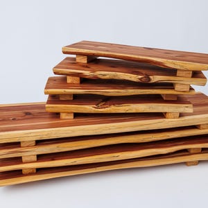 May include: A stack of five wooden serving trays with natural wood grain and a reddish-brown color. The trays are stacked in a staggered pattern, with the largest tray at the bottom and the smallest tray at the top. Each tray has a slightly curved shape and is supported by four small wooden legs.