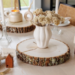 May include: A rustic wooden table setting with two wooden slices, a white vase with dried flowers, and two place cards that say "Welcome".