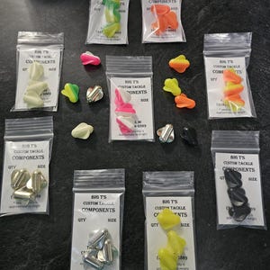 May include: A collection of fishing tackle components in clear plastic bags. The components are various colors, including green, orange, pink, white, and black. Each bag has a label with the text "BIG T'S CUSTOM TACKLE COMPONENTS".