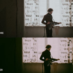May include: A person standing in a room with a window and curtains, holding a black electric guitar. The person is wearing a brown jacket and black pants.