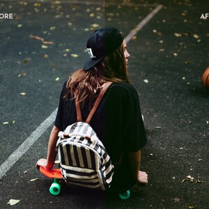 May include: A person wearing a black shirt and a black and white striped backpack sits on a skateboard with a basketball in the background.