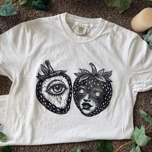 May include: White t-shirt featuring a black and white graphic of two strawberries. One strawberry has an eye design, and the other has a face design. The shirt is short-sleeved and has a crew neck.