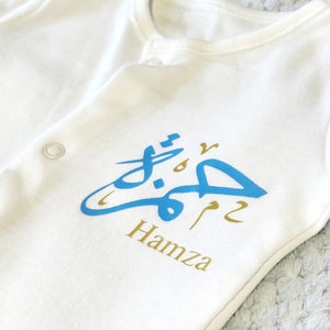 Könnte beinhalten: Weißer Baby-Strampler mit blauer arabischer Kalligraphie und dem Namen "Hamza" in Gold darunter.