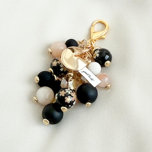 May include: A gold-toned keychain charm featuring a cluster of beads in black, white, and pink. Some beads have a floral design. A small tag reads "good luck!"