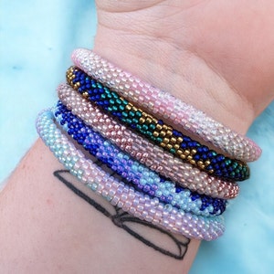 May include: A stack of colorful beaded bracelets in shades of pink, blue, and green. The bracelets are made of small, round beads and are arranged in a layered pattern.