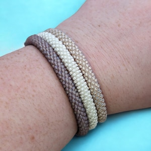May include: A set of four beaded bracelets in shades of beige, white, and gray. The bracelets are stacked on a person's wrist.