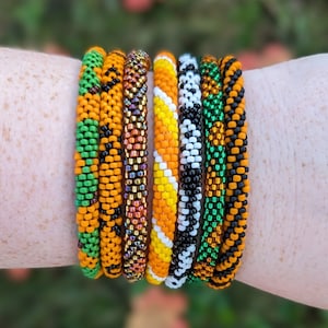 Könnte beinhalten: Ein Stapel bunter Perlenarmbänder in Orange, Grün und Schwarz. Die Armbänder bestehen aus kleinen Perlen, die in verschiedenen Mustern miteinander verwoben sind.