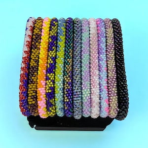 Op de afbeelding: Een verzameling kralenarmbanden in verschillende kleuren, waaronder rood, geel, paars en roze. De armbanden worden gepresenteerd op een zwarte standaard tegen een lichtblauwe achtergrond. Elke armband is gemaakt van kleine, kleurrijke kralen.