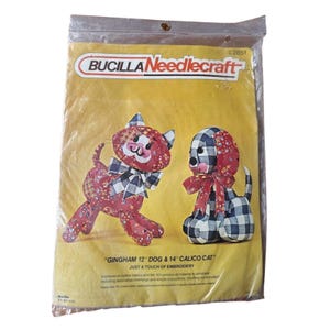 May include: A Bucilla Needlecraft kit in a clear plastic package. The kit features a gingham dog and calico cat design. The text on the package reads "GINGHAM 12" DOG & 14" CALICO CAT". The kit includes materials for embroidery.