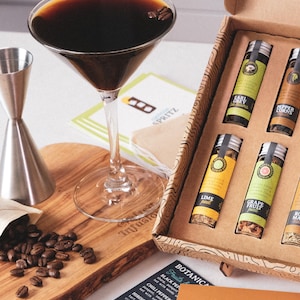 May include: A martini glass with a dark brown cocktail and coffee beans sits on a wooden cutting board. A box of spice infusions and a jigger are nearby. The text "Crafted Infusions" is visible.