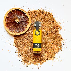 May include: A dried orange slice and a small glass jar of tangerine peel sit on a bed of orange-brown granules. The jar has a yellow label with the text "TANG ERINE peel". The text on the label also reads "light, citrusy & mildly sweet with subtle floral notes".