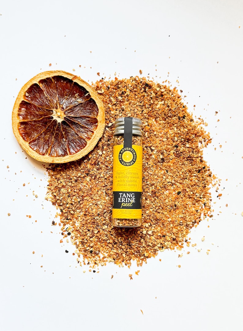 May include: A dried orange slice and a small glass jar of tangerine peel sit on a bed of orange-brown granules. The jar has a yellow label with the text "TANG ERINE peel". The text on the label also reads "light, citrusy & mildly sweet with subtle floral notes".